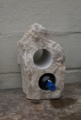 Limestone wine rack with rough texture, cream colored, approximately 14" high with flat cut bottom and a triangular shaped top.  Has two horizontal core holes 3-1/2" diameter cut completely through the stone.  Shown siting atop a table with one bottle of wine in the lower hole.