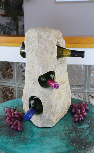 Cream colored Limestone Wine Rack boulder 17 tall, rough textured stone with 3 horizontal holes from top to bottom each offset from the next by 45 degrees.  Showing 3 bottle of wine set atop small table with red grapes.