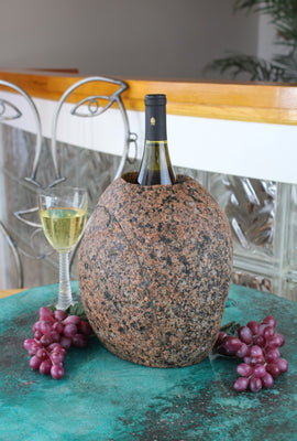 Oval shaped Red-Black Granite Stone Wine Chiller, 10" tall with one  large vertical hole 8" deep and holding one bottle of wine.  Set atop a small table with red grapes and a glass of white wine.