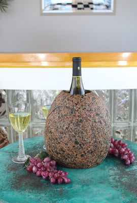 Oval shaped Red-Black Granite Stone Wine Chiller, 10" tall with one  large vertical hole 8" deep and holding one bottle of wine.  Set atop a small table with red grapes and a glass of white wine.