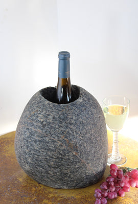 Oval shaped Gray Granite Stone Wine Chiller with one large vertical hole containing one bottle of wine.  Set atop small brown table with red grapes and a glass of white wine.