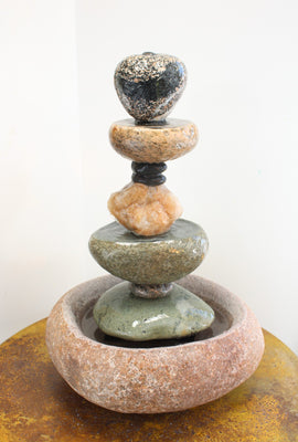 22" tall stone fountain made up of a large circular smooth textured red granite basin stone at the very bottom, filled with water.  A composition of 10 additional stone rising vertically from the basin, made up of mostley smooth textured stones, two of which are caved to create upper pools , a yellow quartz crystal in the middle and a heart shaped top stone.  Fountain is pictured atop a light brown concrete table.