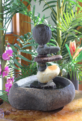 Natural Stone Fountain 19 inches tall consisting of a large smooth hand carved basin stone at the botttom and a asymmetrical composition of nine additional stones some of which are cantilevered to the right, and topped off with a very round shapped top stone.  Background consisting of tropical plants and flowers.