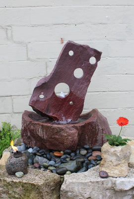 Large Contemporary Purple Quartz stone fountain with multple core hole cut through the upper portion of the composition.  Set atop a basin covered with colorful accent stones and a limestone boulder wall facade.