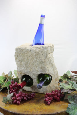 Rectangular cream colored Limestone Wine Rack with one vertical hole at top and two horizontal holes bellow.  Showing blue colored bottle in vertical hole and two horizontal bottles.  All set atop brown table with red grapes and vines.