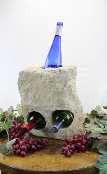 Rectangular cream colored Limestone Wine Rack with one vertical hole at top and two horizontal holes bellow.  Showing blue colored bottle in vertical hole and two horizontal bottles.  All set atop brown table with red grapes and vines.