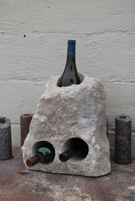 Triangular shaped Limestone wine rack with a white rough texture.  Stone has been drilled to hold 3 bottles of wine, one vertically near the top half of the stone acting as a chiller, and two horizontal side by side near the bottom half.  Bottom of stone has been cut flat.  Shown sitting atop a table  holding 3 bottles of wine.