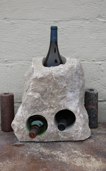 Triangular shaped Limestone wine rack with a white rough texture.  Stone has been drilled to hold 3 bottles of wine, one vertically near the top half of the stone acting as a chiller, and two horizontal side by side near the bottom half.  Bottom of stone has been cut flat.  Shown sitting atop a table  holding 3 bottles of wine.  Stone is approximately 15 inches tall.