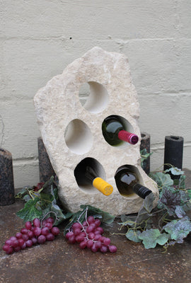 White Limestone wine rack with rough texture. 19 inches tall with 5 horizontal holes cut through the stone to hold wine bottles.  Bottom cut flat and shown sitting atop a table with red grapes and displaying 3 bottles of wine.
