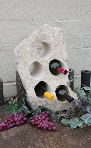 White Limestone wine rack with rough texture. 19 inches tall with 5 horizontal holes cut through the stone to hold wine bottles.  Bottom cut flat and shown sitting atop a table with red grapes and displaying 3 bottles of wine.