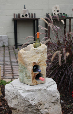 Rough textured Limestone Wine Rack with two vertical holes and two horizontal holes, showing 3 bottles of wine, all set atop a boulder pedestal.  17" tall.  Shown in a outdoor patio display setting.