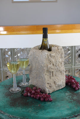 Rectangular shaped rough textured Limestone Wine Chiller 10" tall with one large vertical hole 8" deep holding a bottle of wine.  Set atop a small table with red grapes and two glasses of white wine.