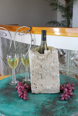 Rectangular shaped rough textured Limestone Wine Chiller 10" tall with one large vertical hole 8" deep holding a bottle of wine.  Set atop a small table with red grapes and two glasses of white wine.