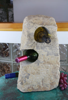 Cream colored Limestone Wine Rack boulder 17 tall, rough textured stone with 3 horizontal holes from top to bottom each offset from the next by 45 degrees.  Showing 3 bottle of wine set atop small table with red grapes.