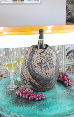 Oval shaped Granite stone wine chiller, 11" tall with one 3-1/2"  diameter hole 8" deep and holding one bottle of wine, set upon table with grapes and two glasses of white wine.