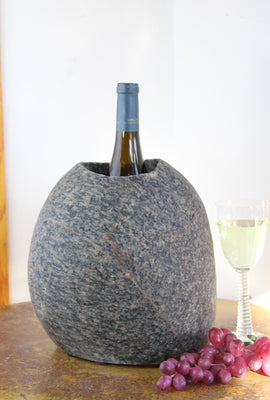 Oval shaped Gray Granite Stone Wine Chiller with one large vertical hole containing one bottle of wine.  Set atop small brown table with red grapes and a glass of white wine.